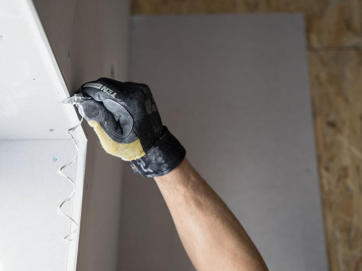 A gloved hand running a taping knife along a drywall corner, smoothing the joint compound.